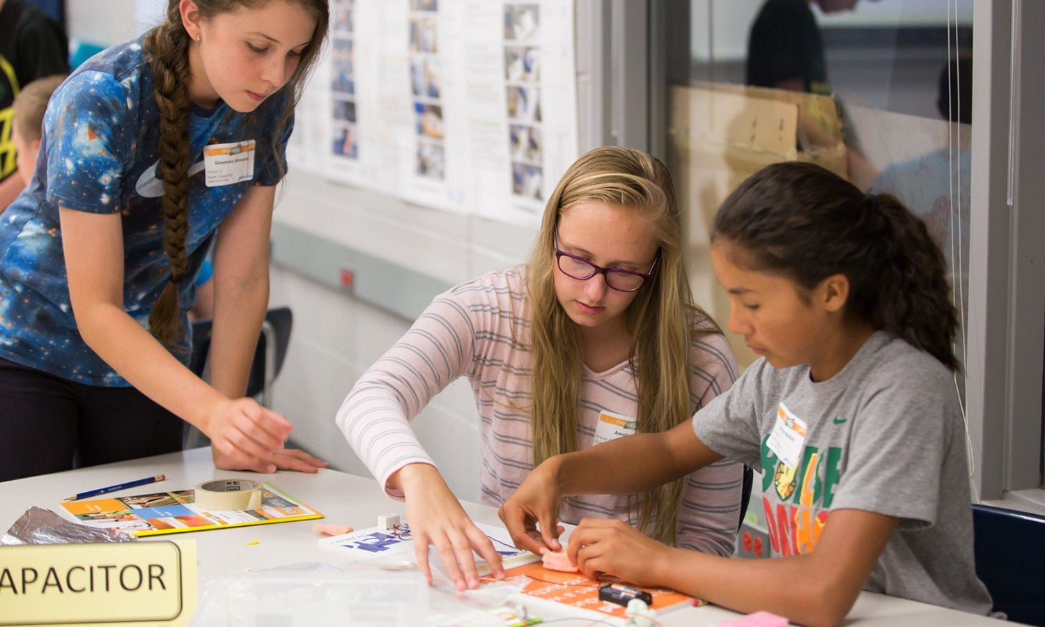Three students collaborate at a table with papers, pens, and an electronic kit. One person stands while two are seated, focused on the task.